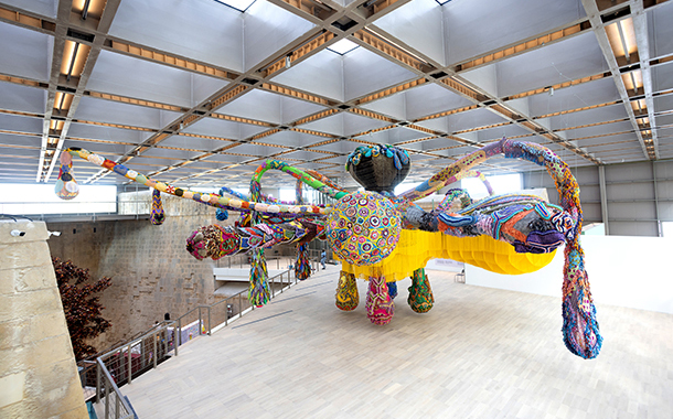 A large, colorful, crocheted sculpture of a creature, hanging from the ceiling of a museum.